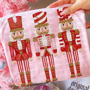May include: A light pink t-shirt featuring three nutcracker figures in red, white, and gold glitter accents. The fabric is printed with a repeating Electric Edge watermark. The nutcrackers wear tall hats and uniforms with gold detailing.
