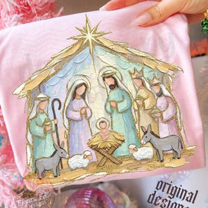 May include: A light pink t-shirt featuring a gold glitter-accented nativity scene illustration. The graphic depicts figures in pastel robes, a manger, a sheep, and a donkey under a star, with the text Electric Edge Designs printed below.