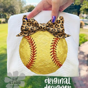 Op de afbeelding: Wit T-shirt met een softbal ontwerp. De softbal is geel met rode stiksels en een luipaardprint strik. De tekst "original designer" is zichtbaar.