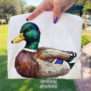 May include: White t-shirt featuring a detailed painting of a mallard duck. The duck has a green head, yellow beak, and brown and gray body. The text "original designer" is printed at the bottom.