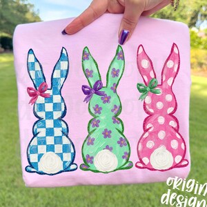 May include: A light pink t-shirt featuring three Easter bunny designs. The bunnies are in blue and white checkered, green with purple flowers, and pink with white polka dots. Each bunny has a bow. The text "original designer" is in the bottom right corner.