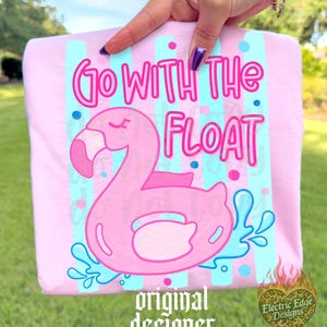 May include: A light pink t-shirt featuring a pink flamingo float with the text "Go With The Float". The design includes blue and pink accents, and the words "original designer" are at the bottom.