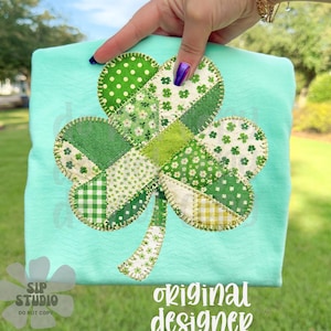 Puede incluir: Una camiseta turquesa clara con un gran diseño de trébol aplicado. El trébol está compuesto por varias telas verdes estampadas, incluyendo lunares, estampados florales y cuadros. El texto "original designer" es visible en escritura blanca.