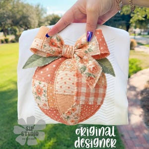 Op de afbeelding: Wit T-shirt met een patchwork pompoen ontwerp en een grote strik. De pompoen is gemaakt van stoffen in oranje, perzik en cr&egrave;me, met bloemen- en stippenpatronen. De strik heeft een bloemenpatroon. De tekst "original designer" is zichtbaar.