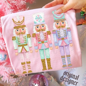 May include: A light pink t-shirt featuring a graphic print of three nutcracker figures in pastel colours with gold glitter accents. The nutcrackers are dressed in uniforms with hats, boots, and intricate detailing. The text original designer is printed in black in the bottom right corner.