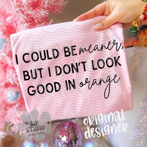 May include: A light pink t-shirt with the text "I COULD BE meaner, BUT I DON'T LOOK GOOD IN orange" in black script. The shirt has a zig-zag pattern and is held up by a hand. The background includes a pink Christmas tree and disco balls.