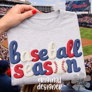 May include: A light gray t-shirt with the words "Baseball Season" in blue, red, and white, with a baseball graphic. The shirt is held up in front of a baseball stadium.