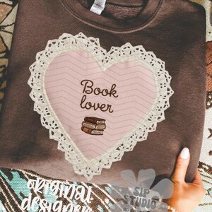 May include: A brown sweatshirt featuring a pink heart design with the words "Book lover" and a stack of books. The heart is trimmed with white lace. The sweatshirt is held by a person.