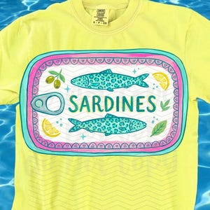 May include: A yellow t-shirt with a graphic of a sardine can. The can design features two turquoise fish with leopard print patterns, olives, lemon slices, and the word "SARDINES". The shirt has the text "original designer" at the bottom.
