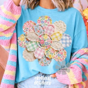 Op de afbeelding: Lichtblauw T-shirt met een bloemen patchwork design. Het shirt heeft de tekst "original designer" erop gedrukt. Een kleurrijke gestreepte trui is over de schouders gedrapeerd. De afbeelding heeft een lente- of Paasthema.