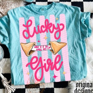 May include: A turquoise t-shirt featuring a pink design with the words "Lucky Girl." The design includes a fortune cookie with the text "What if it all works out?" and pink vertical stripes. The shirt has short sleeves.
