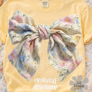 May include: A yellow t-shirt featuring a large bow design with a floral pattern in pink, blue, and yellow. The text "original designer" is printed in white. The shirt has rolled-up sleeves.