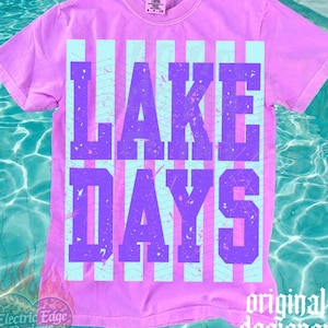 May include: A lilac-colored t-shirt with the words "LAKE DAYS" in purple block letters. The text is set against a backdrop of light blue vertical stripes. The shirt has a crew neck and short sleeves. The words "original designer" are printed on the bottom right.