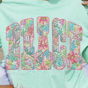 May include: A light green t-shirt with the word "BEACH" in large, colorful letters. The letters are filled with a floral and aquatic pattern, including seahorses and starfish. The shirt is a relaxed fit.