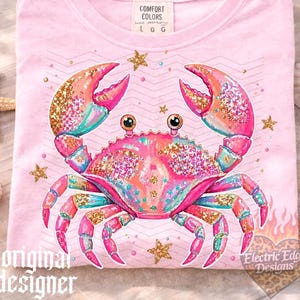 May include: Pink t-shirt featuring a colorful crab design with glitter accents and star details. The crab has pink, orange, and teal hues. The shirt includes the text "original designer" and "Electric Edge Designs".