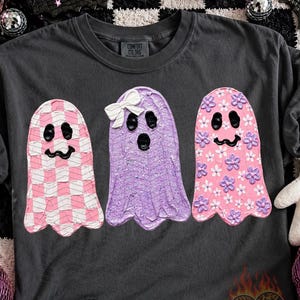May include: A charcoal gray t-shirt featuring three ghost illustrations. The left ghost has a pink and white checkered pattern, the middle ghost is solid purple with a white bow, and the right ghost is pink with small purple and white flower accents.