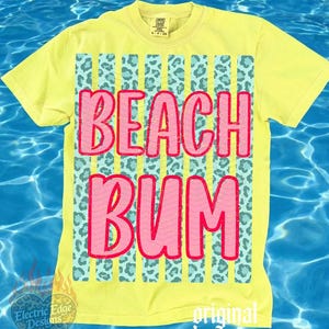 May include: Yellow t-shirt with the words "BEACH BUM" in pink, outlined in red. The text is set against a background of vertical stripes with a teal leopard print. The shirt has short sleeves and a crew neck.