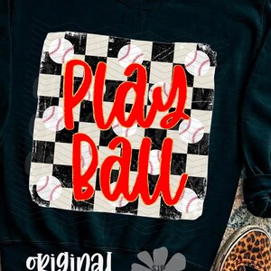 May include: Black sweatshirt with a baseball-themed design. The design features a checkered pattern with baseballs and the words "Play Ball" in red. The words "original designer" are printed at the bottom.