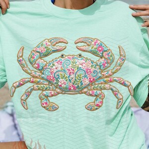K&ouml;nnte beinhalten: Hellgr&uuml;nes T-Shirt mit detailliertem Krabben-Design. Die Krabbe ist mit einem Blumenmuster in Rosa, Blau und Gold verziert. Das T-Shirt hat kurze &Auml;rmel und eine lockere Passform, ideal f&uuml;r einen Tag am Strand.