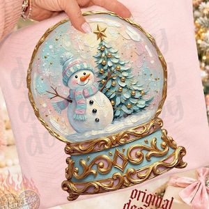 May include: A light pink sweatshirt featuring a graphic of a snow globe with a snowman wearing a blue and pink striped hat and scarf, standing next to a decorated Christmas tree with a gold star, set on an ornate gold and blue base.