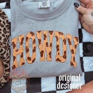 May include: A gray sweatshirt with the word "HOWDY" in a brown, textured font. The sweatshirt is on a black and white checkered blanket. A leopard print slipper is visible on the left.