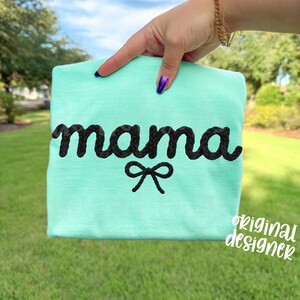 May include: A light teal t-shirt with the word "mama" embroidered in black, braided-style lettering. A small black bow is embroidered below the word. The words "original designer" are written in white script in the lower right corner.
