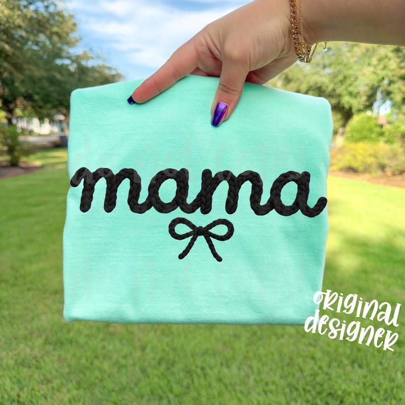 May include: A light teal t-shirt with the word "mama" embroidered in black, braided-style lettering. A small black bow is embroidered below the word. The words "original designer" are written in white script in the lower right corner.