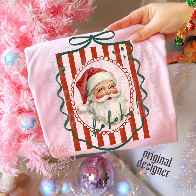 May include: A light pink t-shirt featuring a graphic of Santa Claus with a white beard and red hat, centered within a red and white striped rectangular frame. The design includes a dark green scalloped border and the handwritten text ho ho ho.