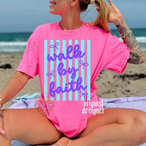 May include: A bright pink t-shirt with the words "walk by faith" in a cursive font, set against a vertical striped background of light blue. The shirt has a crew neck and short sleeves. The words "original designer" are printed on the shirt.