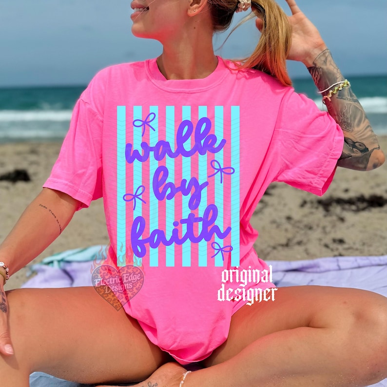 May include: A bright pink t-shirt with the words "walk by faith" in a cursive font, set against a vertical striped background of light blue. The shirt has a crew neck and short sleeves. The words "original designer" are printed on the shirt.