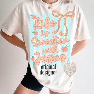 May include: Cream-colored t-shirt with the phrase "Life is Sweeter with Jesus" in peach-colored script. The design includes peach illustrations and a peach-colored bow. The words "original designer" are printed below the main text.