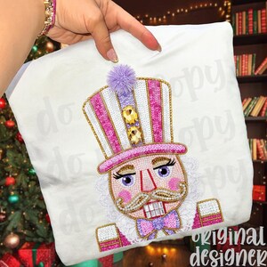 May include: White pillow with an embroidered design of a nutcracker. The nutcracker has a pink and white striped hat, a gold mustache, and a purple bow tie. The design includes sequins and a fluffy purple pom-pom.