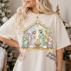 May include: A cream-colored short-sleeve t-shirt featuring a colorful graphic of a nativity scene. The illustration depicts the holy family, wise men, and animals under a golden star. The text original designer is printed in black on the lower right.