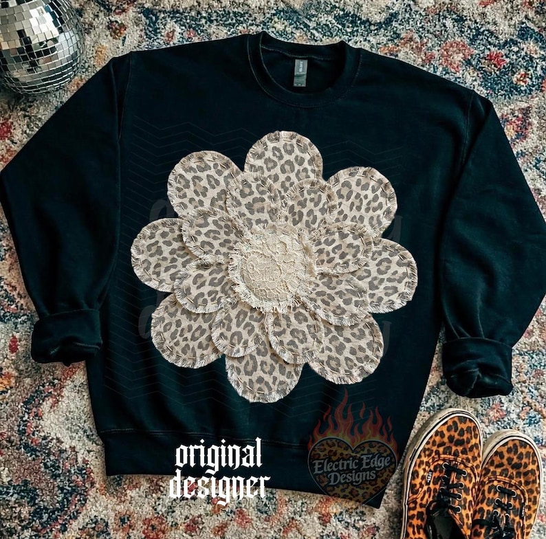 May include: Black sweatshirt with a large, layered flower design. The flower petals feature a leopard print pattern, and the center has a cream-colored, textured fabric. The words "original designer" are printed on the bottom. Leopard print shoes are in the bottom right corner.