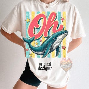 May include: Cream-colored t-shirt featuring a blue whale illustration with the word "Oh" in red script. The design includes yellow stars and vertical stripes. The text "original designer" is printed below the whale.