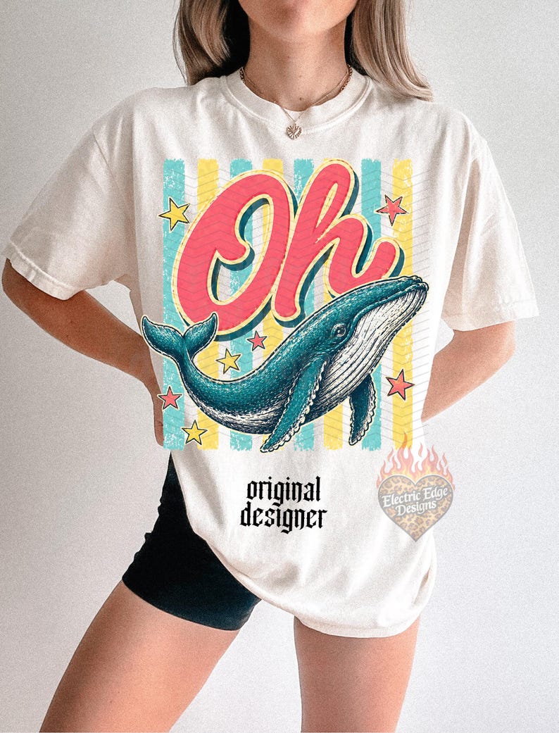 May include: Cream-colored t-shirt featuring a blue whale illustration with the word "Oh" in red script. The design includes yellow stars and vertical stripes. The text "original designer" is printed below the whale.