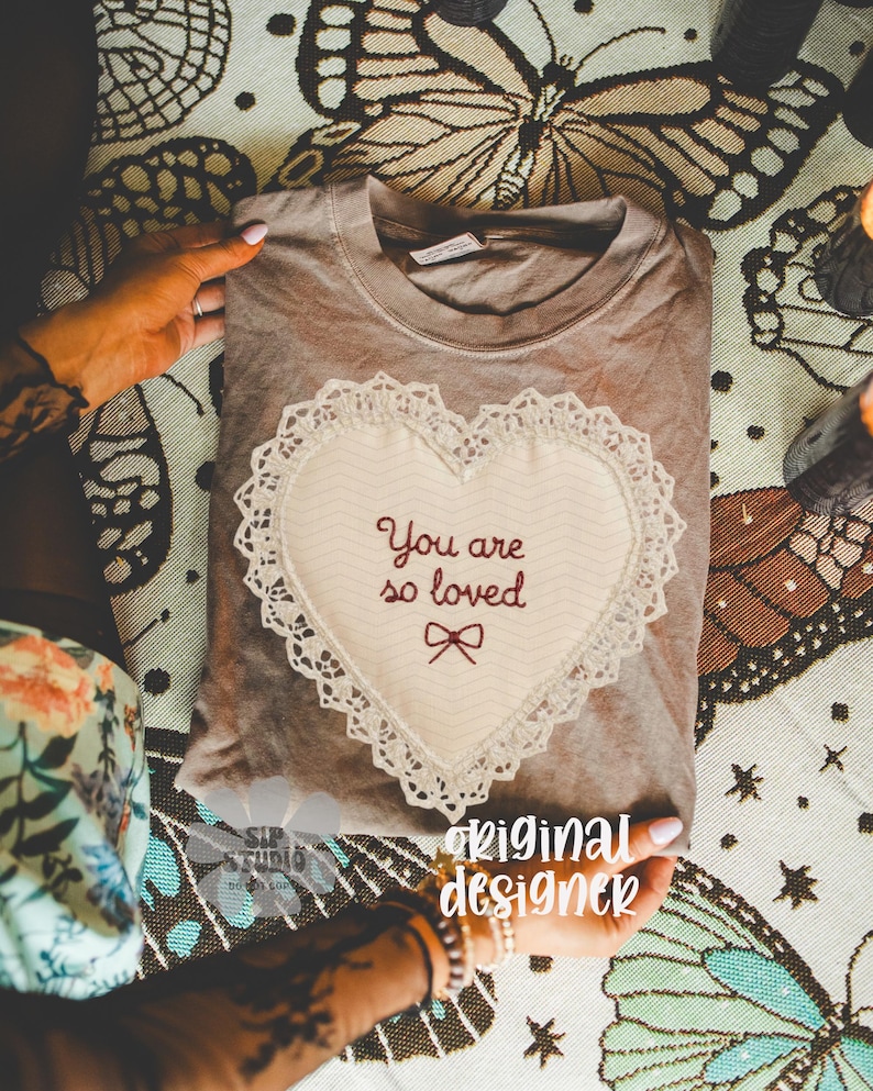 May include: A taupe t-shirt with a heart-shaped appliqu&eacute; embroidered with the words "You are so loved" and a bow. The heart is trimmed with lace. The shirt is folded and displayed on a patterned surface, with the words "original designer" visible.