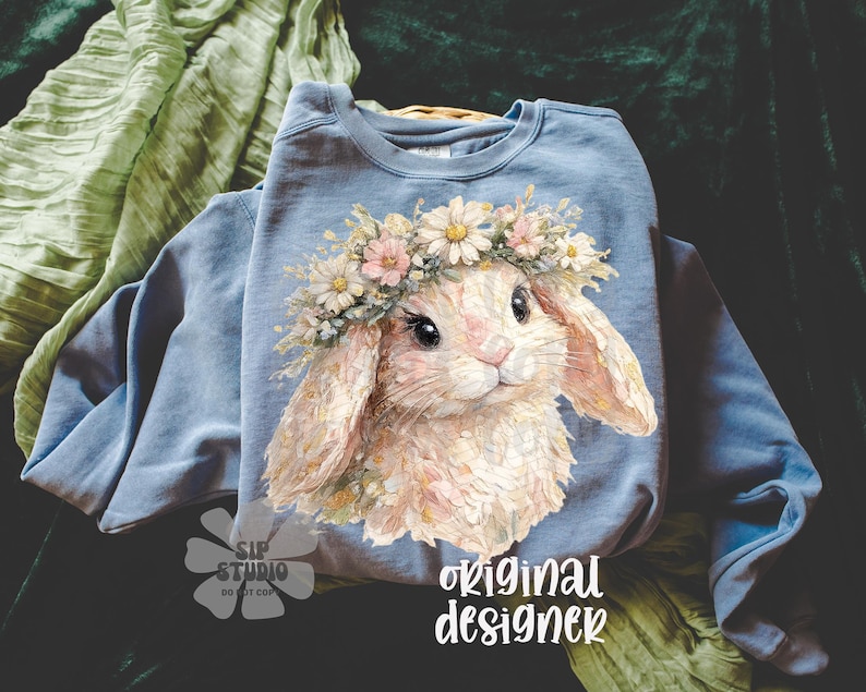 May include: A light blue sweatshirt featuring a watercolor illustration of a bunny wearing a flower crown. The bunny has soft, pastel colors and is centered on the shirt. The words "original designer" are printed in white script.