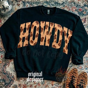 May include: Black sweatshirt with the word "HOWDY" in a tan and brown cowhide print. The sweatshirt has long sleeves and a crew neck. The words "do not copy" and "original designer" are printed on the sweatshirt. Leopard print shoes are at the bottom.