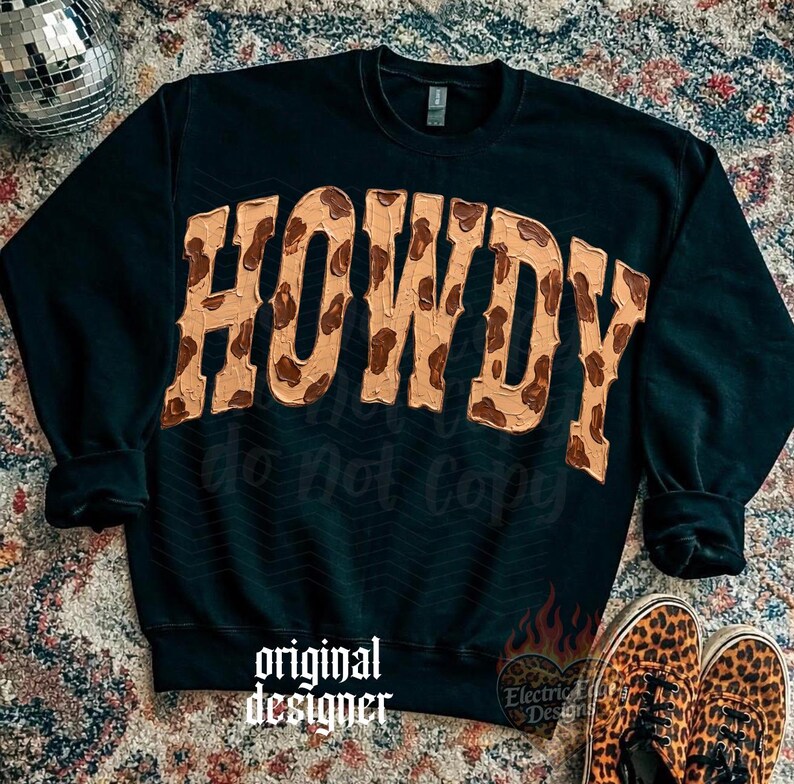May include: Black sweatshirt with the word "HOWDY" in a tan and brown cowhide print. The sweatshirt has long sleeves and a crew neck. The words "do not copy" and "original designer" are printed on the sweatshirt. Leopard print shoes are at the bottom.