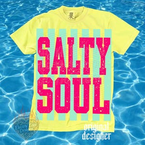 May include: A yellow t-shirt with the words "SALTY SOUL" in large, distressed pink letters. The text is set against a background of vertical stripes in light blue and yellow. The shirt has a crew neck and short sleeves.