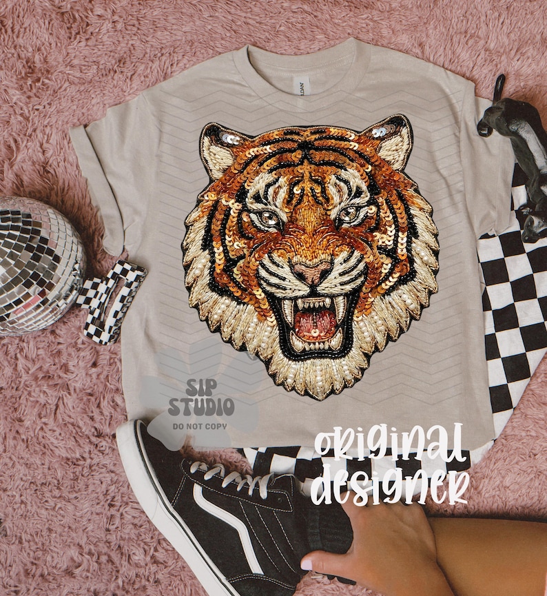 May include: Beige t-shirt featuring a detailed tiger face design. The tiger's face is embellished with sequins in shades of gold, orange, and black. The shirt is paired with black and white checkered accessories and a black sneaker.