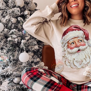 Puede incluir: Una sudadera beige de cuello redondo con un gr&aacute;fico de pintura al &oacute;leo texturizada estilo impasto de Santa Claus con gorro rojo y barba blanca. La sudadera tiene un sutil patr&oacute;n de chevr&oacute;n y el texto "original designer" en letra cursiva.