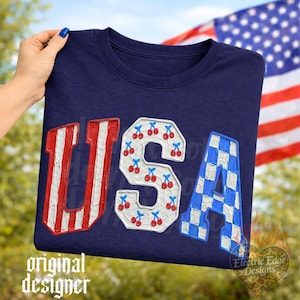 May include: Navy blue crew neck sweatshirt with the word "USA" in red and white striped, cherry-filled, and blue and white checkered letters. The American flag is in the background.