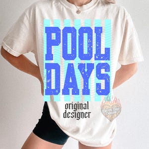 May include: Cream-coloured t-shirt featuring the words "POOL DAYS" in large blue letters, set against a light blue wavy pattern. The text "original designer" is below in a gothic font.