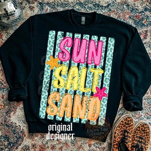 May include: Black sweatshirt with the words "SUN SALT SAND" in pink, yellow, and orange, with starfish accents. The design is on a background of vertical stripes and leopard print. The words "original designer" and "Electric Edge Designs" are also printed on the sweatshirt.