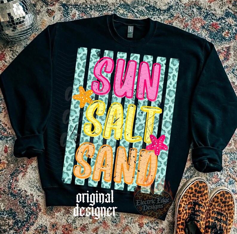 May include: Black sweatshirt with the words "SUN SALT SAND" in pink, yellow, and orange, with starfish accents. The design is on a background of vertical stripes and leopard print. The words "original designer" and "Electric Edge Designs" are also printed on the sweatshirt.