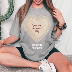 May include: A grey t-shirt with a heart-shaped design and the text "You are so loved." The heart has a cream-coloured centre with a lace border and a small bow. The words "original designer" are printed below the heart.