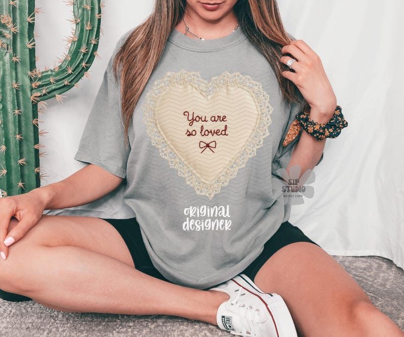 May include: A grey t-shirt with a heart-shaped design and the text "You are so loved." The heart has a cream-coloured centre with a lace border and a small bow. The words "original designer" are printed below the heart.