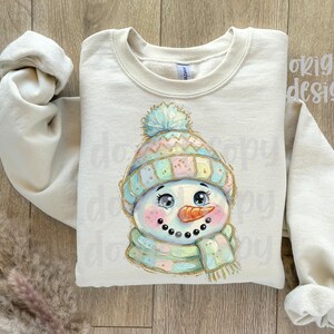 May include: A cream-colored crewneck sweatshirt featuring a central graphic of a snowman wearing a pastel-colored knit hat with a pom-pom and a matching scarf. The snowman has a bright orange carrot nose, rosy cheeks, and large dark eyes.